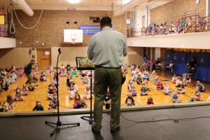 Principal standing on stage reading to children sitting on the gym floor below