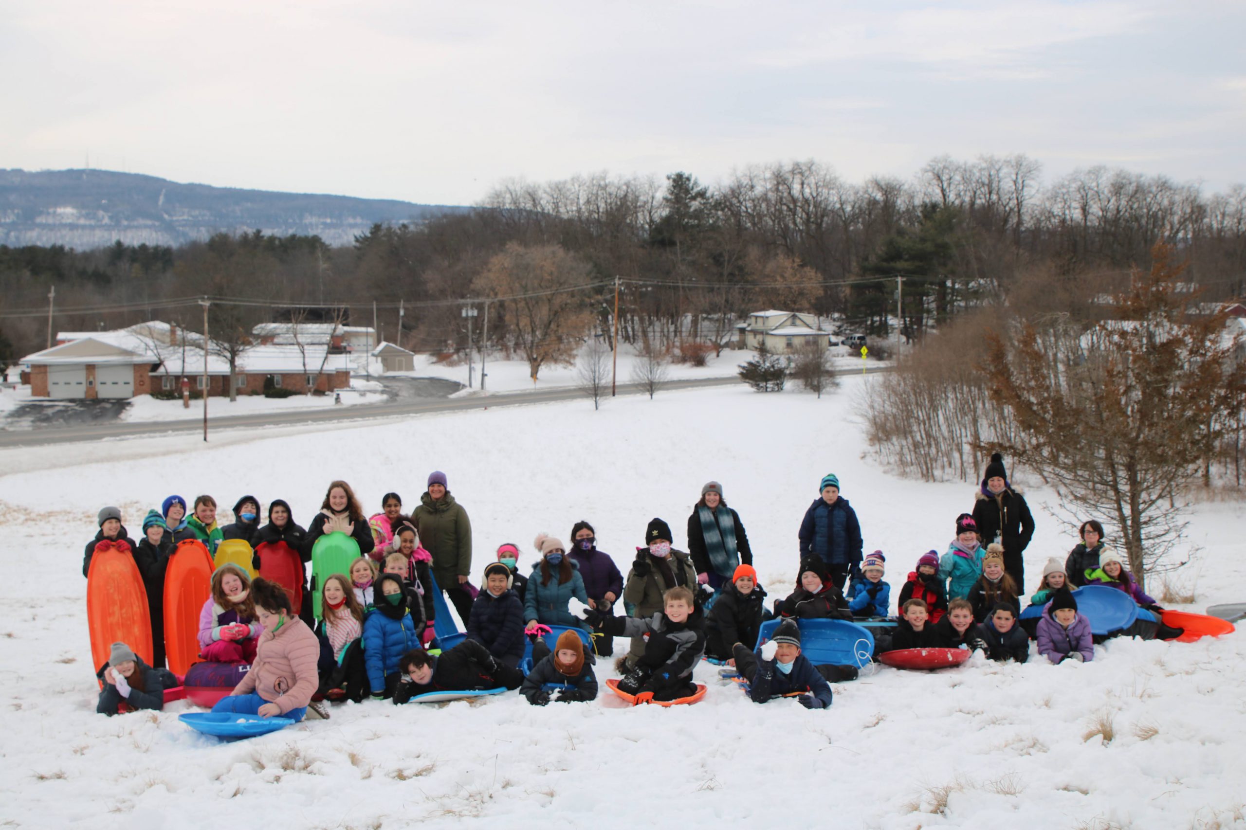 fifth grade class standing together outside in the snow. All are wearing snow gear and some have sleds and tubes.