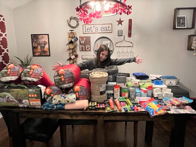 student with her arms wide across a table of suitcases and medical supplies and other necessities for Ukraine