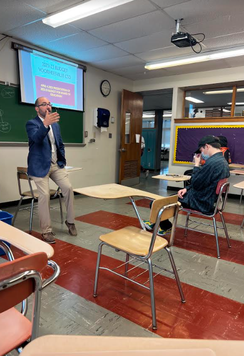 teacher siting on desk in front of classroom with a projector screen behind him