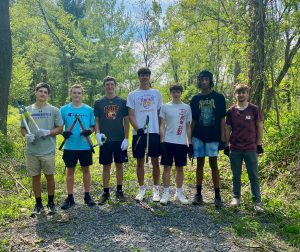 seven students standing in a row at a park each are holding clippers and other yard work tools