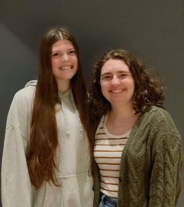 a student with long brown hair wearing a gray sweatshirt and a student with shoulder length curly hair wearing a green sweater and stripped shirt
