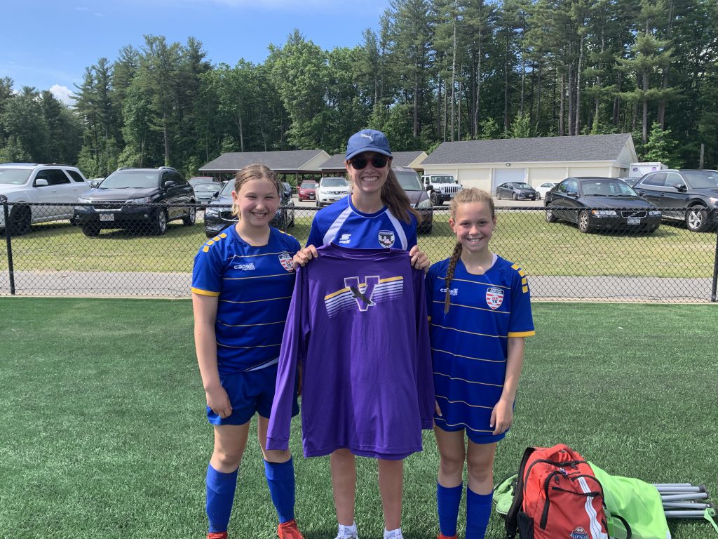 two young soccer players in blue uniforms stand on each side of the coach who is holding up a purple Voorheesville sweatshirt