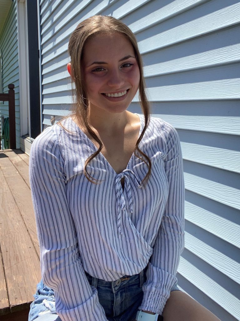 student with long brown hair smiles outside on a deck wearinf a blue and white striped shirt