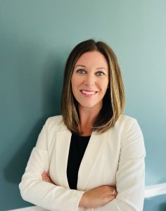 woman wearing white jacket and black shirt stands with her arms crossed against a sage green wall