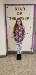 Star of the week Ava stands in front of bulletin board. She is smiling toward the camera.