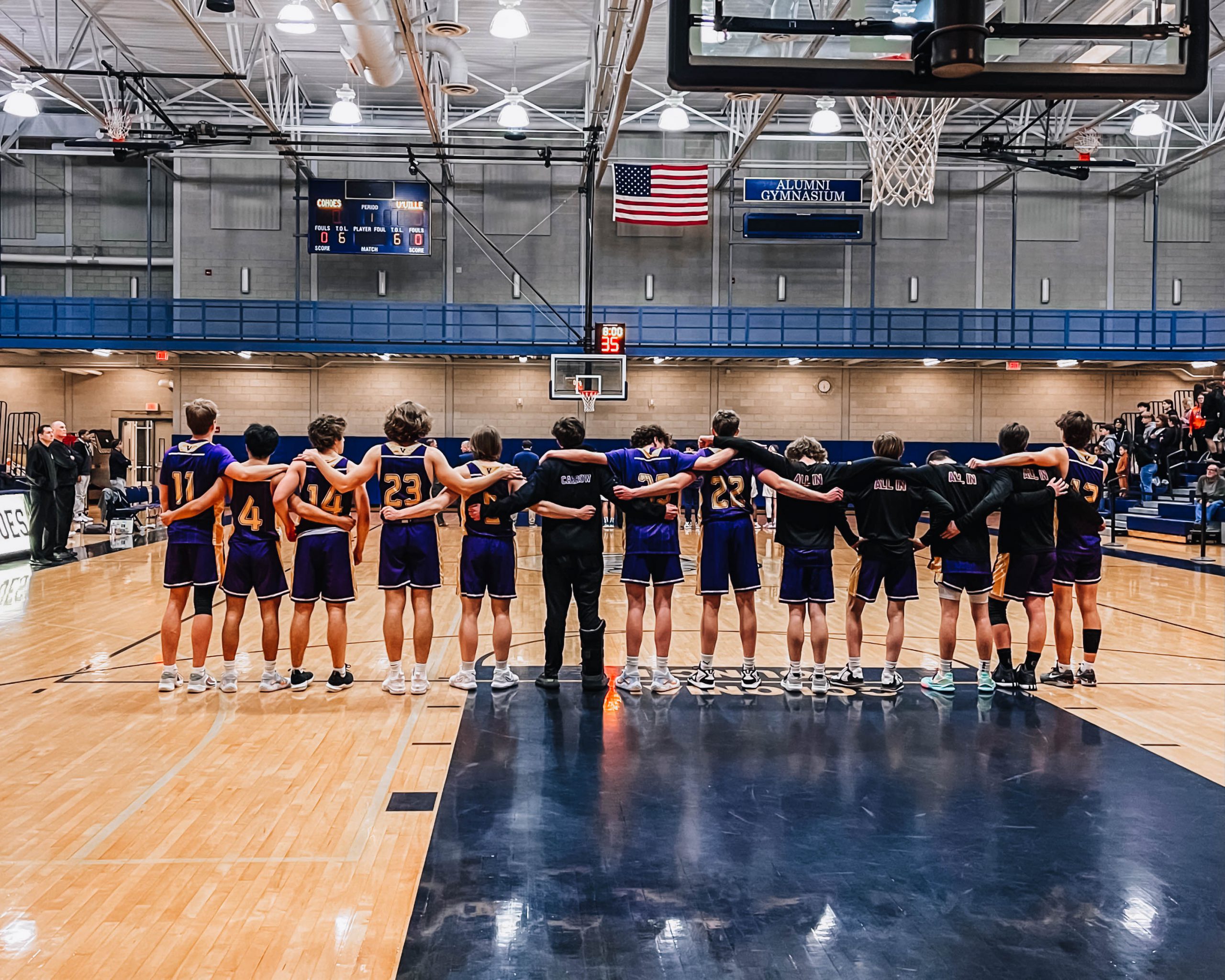Members of the Voorheesville Basketball Team stand in a line with their arms around each other's shoulders. They are on the court, facing away from the camera.