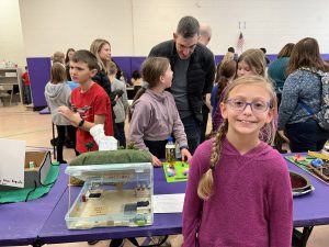 A 4th grade student smiles as she stands in front of her project for mitigating natural disasters. A crowd of people is looking at other projects that are displayed in the room.