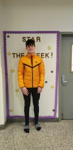 Student stands in front of Star of the Week bulletin board. He is smiling.