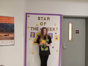 Girl stands in front of Star of the Week bulletin board. She is smiling.