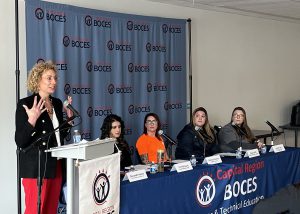 Woman stands at podium. 4 women are seated at a table to her left.