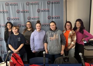 A group of teen girls stand in a group with a teacher. In the background is a Capital Region BOCES banner.