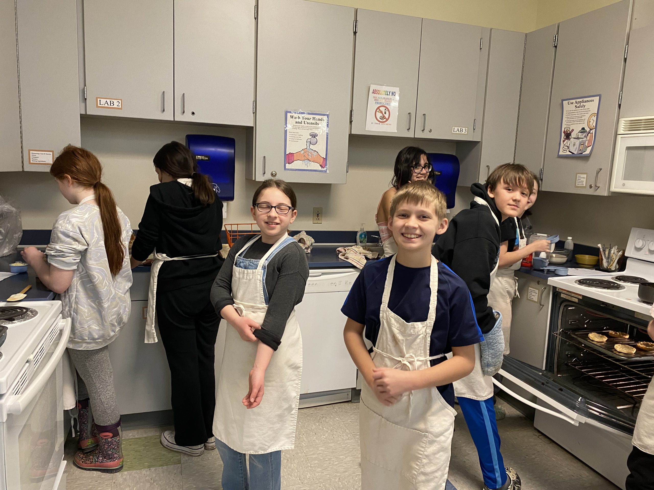 Group o middle school students wearing aprons prepare food in a kitchen.
