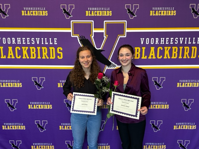 Two teens stand in front of Voorheesville Blackbirds banner. They are each holding a certificate and a rose.