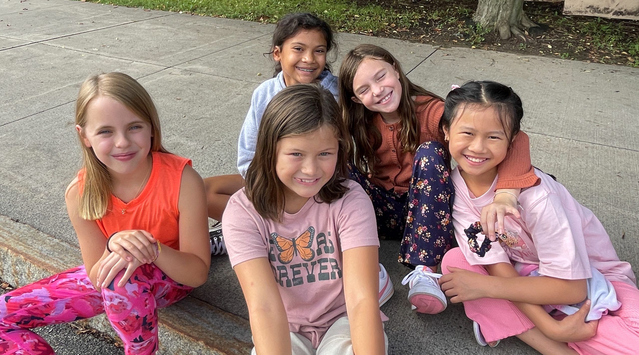 Five elementary students sit on sidewalk.
