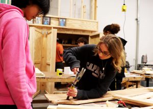 Emily Grace Michaud uses a hammer and awl on wood. Other students are in the background.