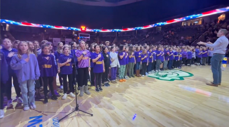 Wearing purple, the middle school choir stands with their hands over their hearts while they sing in arena.