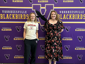 VCSD Class of 2025 salutatorian, Lindsey Wight and valedictorian Brianna Hillmann, stand in front of a bright purple backdrop covered with the Voorheesville logo and a large V. Both are looking at and smiling for the camera.