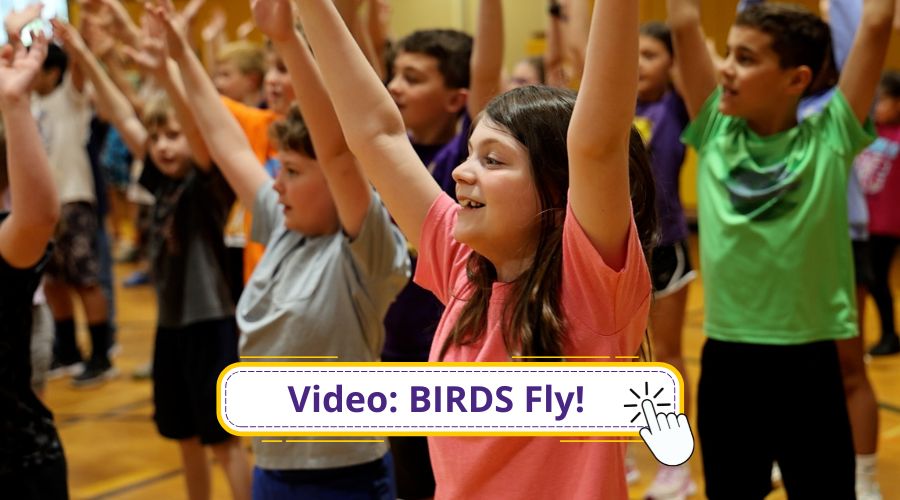 Large group of grade four students in gym with their hands raised above their heads. Watch our BIRDS video. Click here hand.