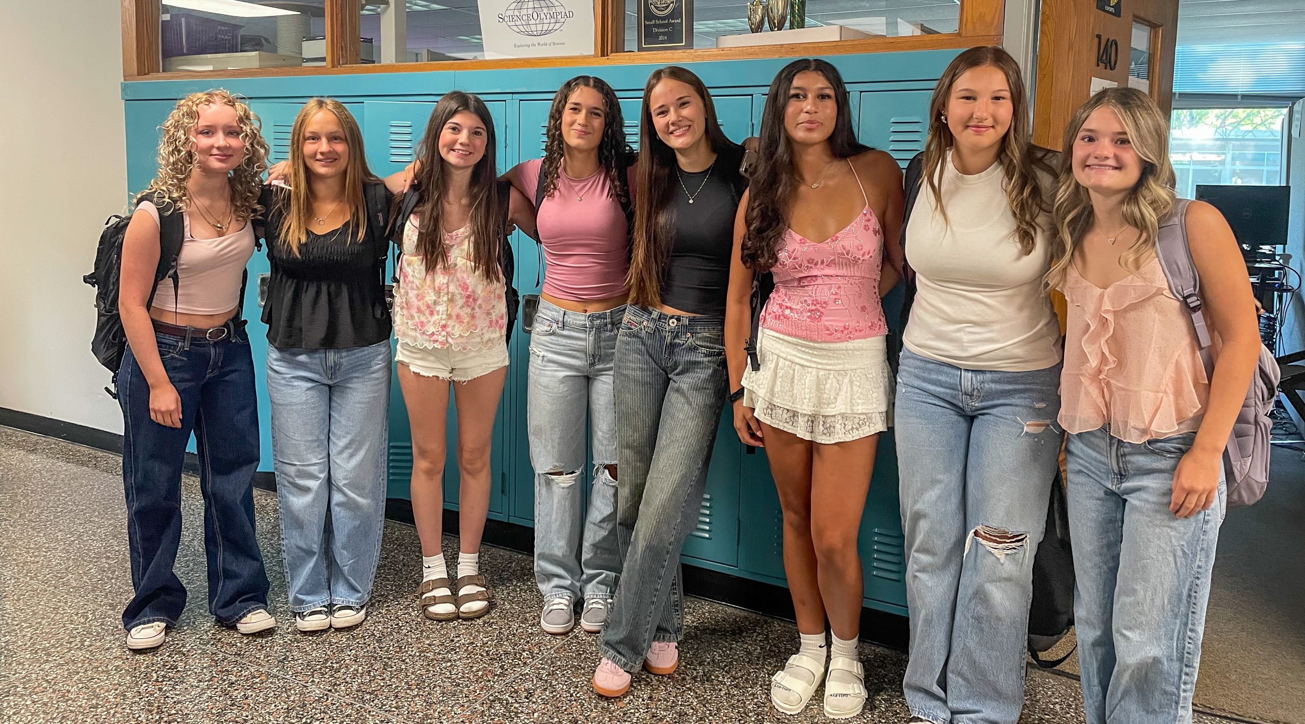 Eight high school students stand together, some with arms on each other's shoulders, in front of lockers.