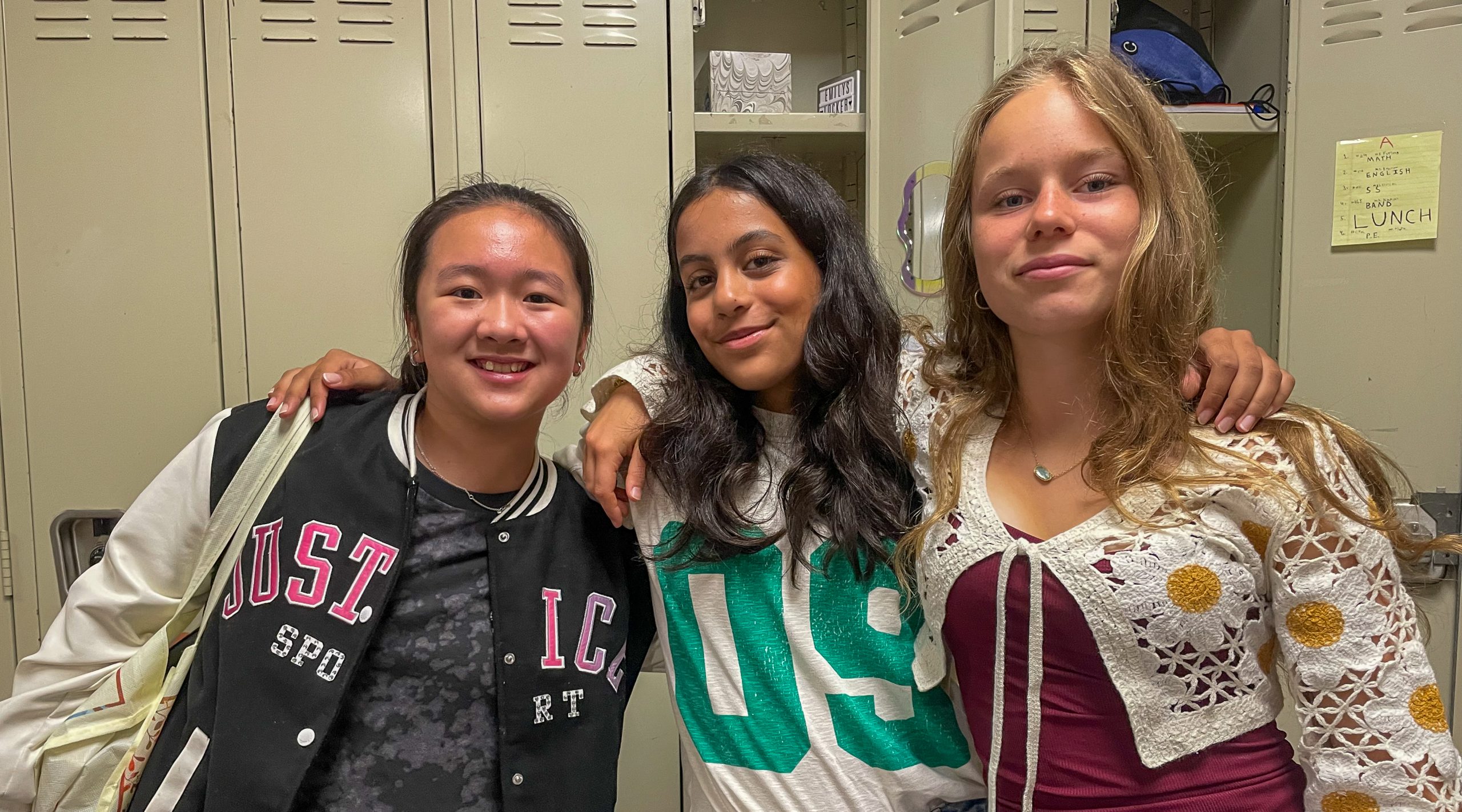 Three middle school students stand with arms around each other's shoulders in front of locker.