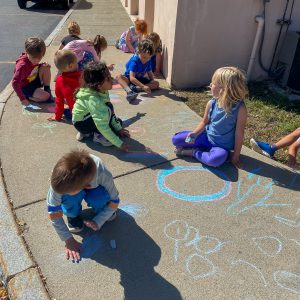 A group of kindergarten students use chalk on a sidewalk outside elementary school.