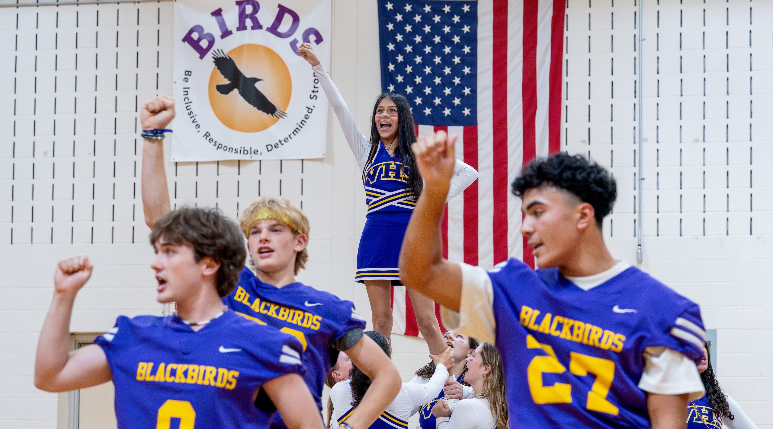 Football players and cheerleader perform together at pep rally. BIRDS poster and American flag in the background.