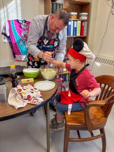Boy, wearing a chef's hat, makes pancakes with Principal Jeff Vivenzio in his office. Vivenzio is wearing a chef's apron.