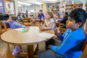 Students sit around round tables in a school library, holding pencils and working on a classroom activity while a teacher leads the group. Bookshelves and classroom materials are visible in the background.