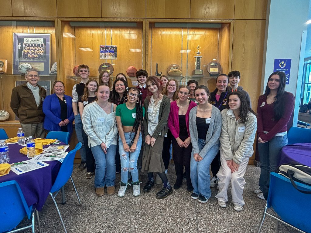 A large group of students and adults stands together for a group photo in front of a wooden display case filled with sports trophies and memorabilia. Tables with purple tablecloths and chairs are visible on either side of the group.