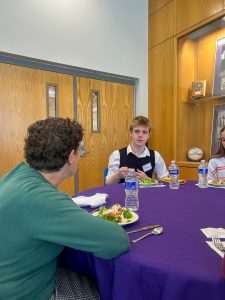 Two people sit across from each other at a table with salad and water bottles, talking near wooden double doors.