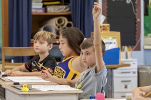 Three elementary students at desks. One has their hand raised.