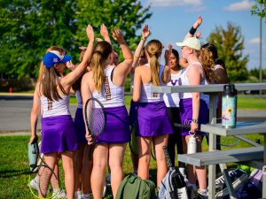 Members of tennis team huddle and raise their right hands. 