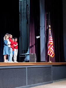 Elementary school students stand on stage facing the U.S. flag while reciting the Pledge of Allegiance.