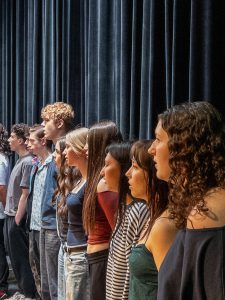 A group of students stands in rows on stage, singing during a ceremony.