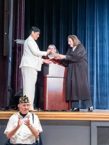 A newly naturalized citizen holds a certificate of citizenship while standing on stage near a lectern during a naturalization ceremony.