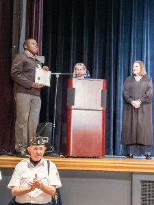 A newly naturalized citizen holds a certificate of citizenship while standing on stage near a lectern during a naturalization ceremony.